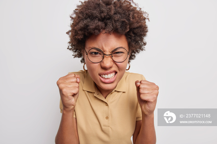 Afro American woman clenches teeth and fists angrily being irritated with something cannot stand annoying noise wears spectacles casual t shirt isolated over grey background. Negative emotions