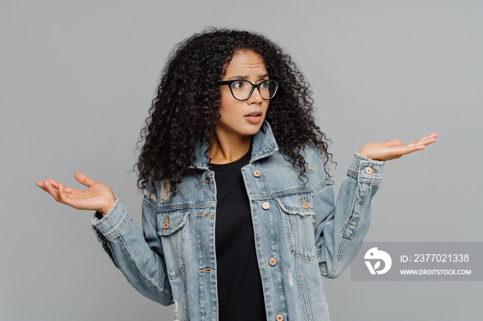 Confused unaware Afro woman with crisp hair, raises hands in bewilderment, looks aside, cannot make decision, wears spectacles and jean jacket, isolated over grey background. What should I do now?