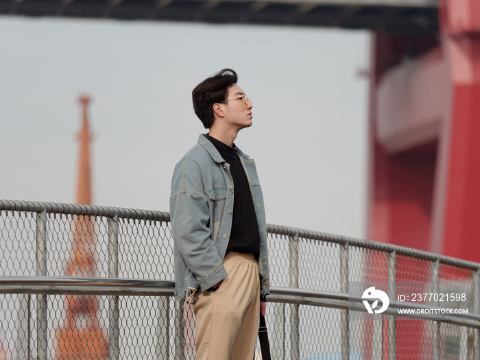 Portrait of a handsome Chinese young man standing and looking away  with hand in pocket in windy sunny day, has nothing to do just waiting.