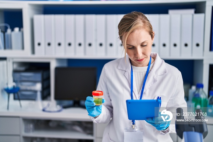 Young blonde woman scientist using touchpad holding urine test tube at laboratory