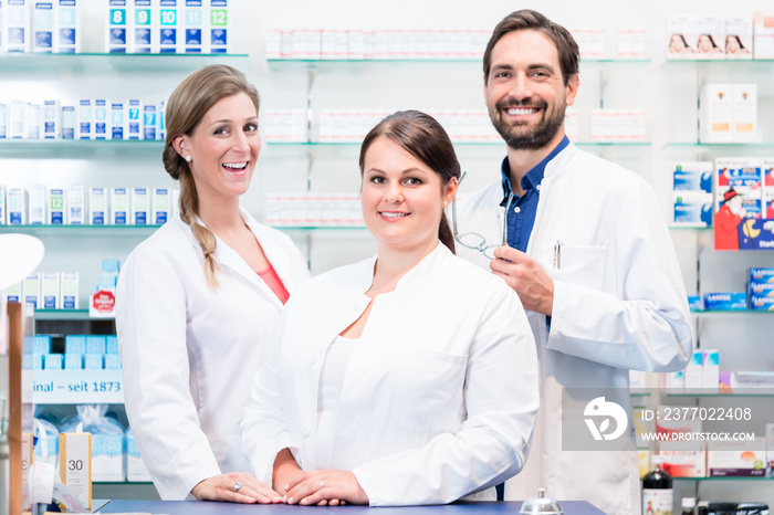 Team of apothecaries in pharmacy standing in front of shelf with prescription drugs