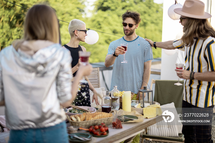 Young group of stylish people having a festive dinner on the roof terrace. Hipsters hanging out together, drinking wine and talking outdoors