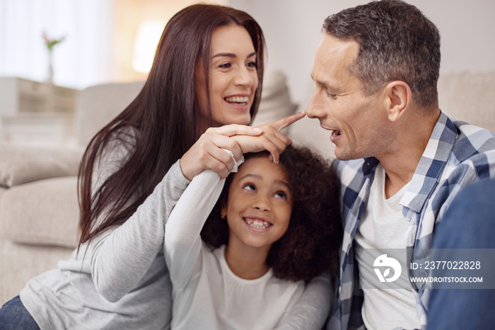 Fooling around. Pretty content young long-haired woman smiling and sitting on the floor with her husband and daughter and they touching the man on the nose