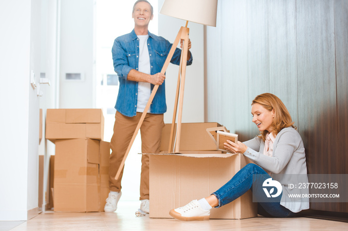 smiling man holding floor lamp while woman sitting in floor and unpacking carton box