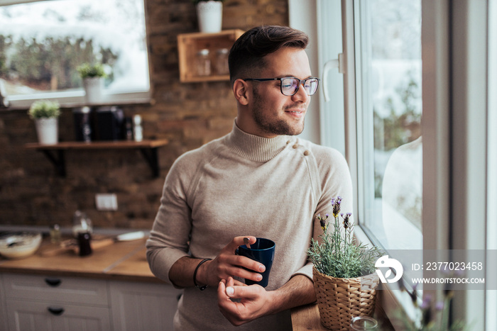 Handsome young man looking through at cozy home and drinking tea or coffee.