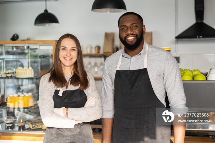 Smiling caucasian waitress and cheerful African-American waiter stands with arms crossed behind the counter. Confident multiracial baristas look at the camera