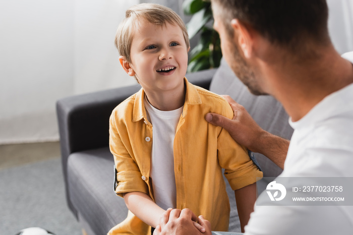 selective focus of father touching shoulder and holding hand of smiling son while talking to him at home