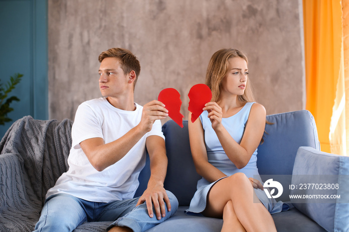 Young couple holding halves of broken heart on sofa at home. Relationship problems