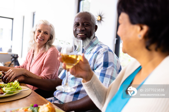 Multiracial senior couple holding hands while woman enjoying wine at dining table in nursing home