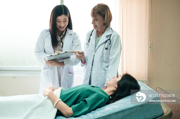 Caucasian senior woman doctor teach Asian medical student by bedside teaching technique to diagnosis patient in the ward at hospital. Working together of doctors of different ethnicity.