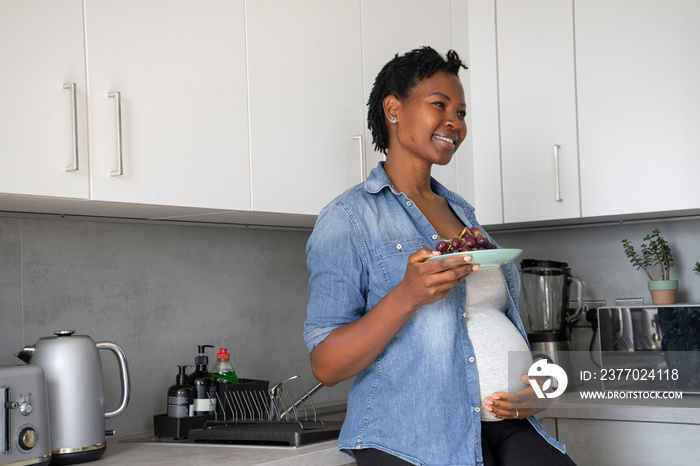 Smiling pregnant woman holding plate with red grapes in kitchen
