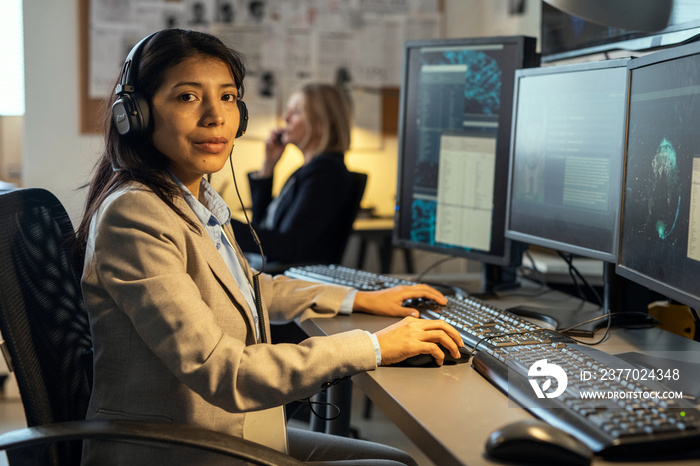 Young confident Hispanic female agent of secret service with headphones looking at camera while working in office