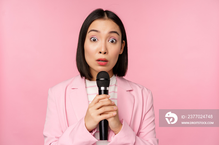 Young asian saleswoman, office lady with suit, holding microphone and looking shocked at camera, talking, giving speech, standing over pink background