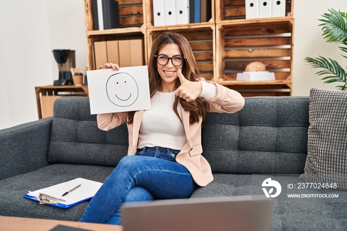 Young brunette woman on therapy session working at consultation office smiling happy and positive, thumb up doing excellent and approval sign