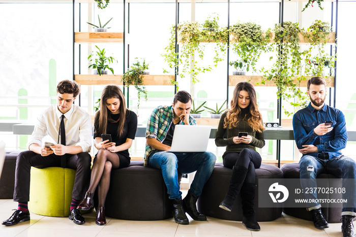 Group of friends sitting on chairs near each other and everyone use his divices in modern office room. Together fun in device.