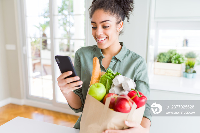 Young african american girl holding paper bag of groceries and using smartphone with a happy face standing and smiling with a confident smile showing teeth