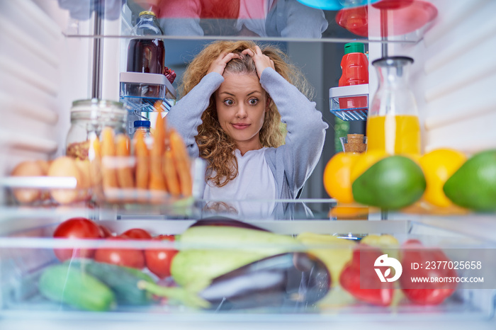Confused woman choosing what to eat. Hands on head. Picture taken from the inside of fridge full of groceries.