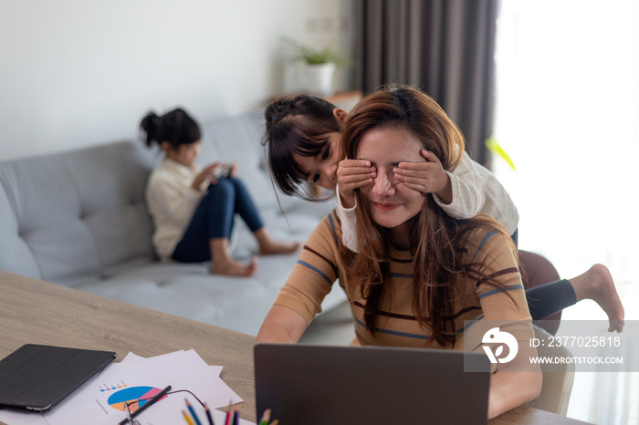 woman sits by the table at home office during lockdown, working on laptop. Playful child distracts from work, covering her mother’s eyes, kid making noise and asking attention from busy mom