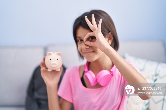 Young girl holding piggy bank smiling happy doing ok sign with hand on eye looking through fingers