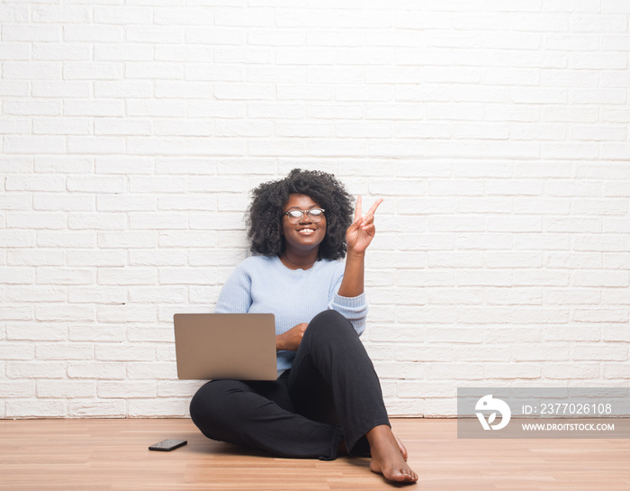 Young african american woman sitting on the floor using laptop at home smiling with happy face winking at the camera doing victory sign. Number two.