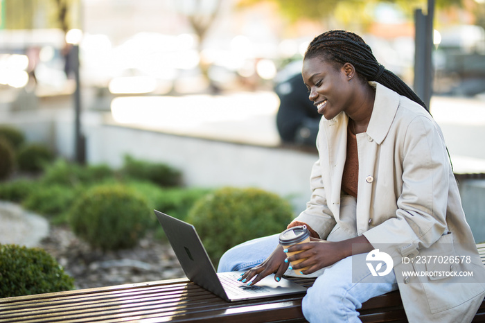 Young african woman in park. Beautiful woman sitting on bench drinking coffee and using laptop via airpods.