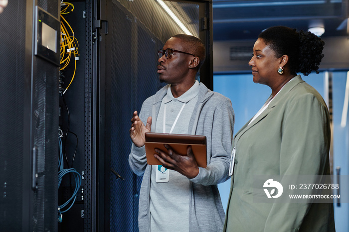 Waist up portrait of black IT engineer with female assistant in server room inspecting network systems