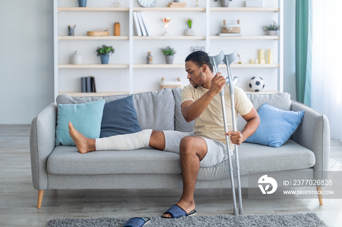 Young African American guy with plastered leg sitting on sofa, leaning on crutches at home