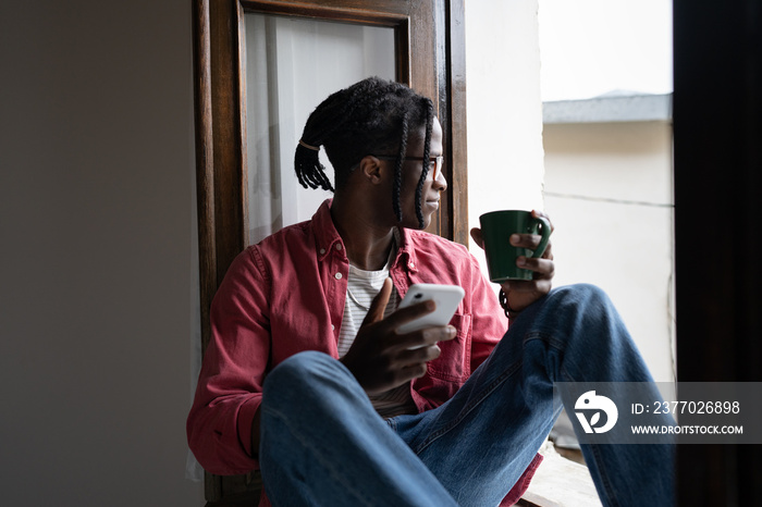 Young smiling African guy holding smartphone and cup of tea enjoying lazy weekend day at home, sitting on windowsill and looking out window. Calm black man turning off phone to spend time in silence