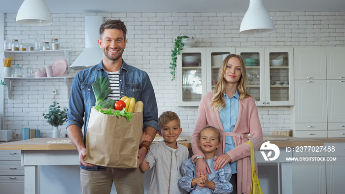 Smiling man holding paper bag with fresh food near family with kids in kitchen