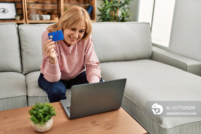 Middle age blonde woman buying using laptop and credit card sitting on the sofa at home.