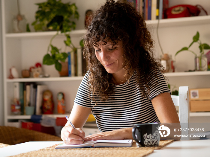 Caucasian woman with curly hair writing in a notebook sitting in the living room of an apartment