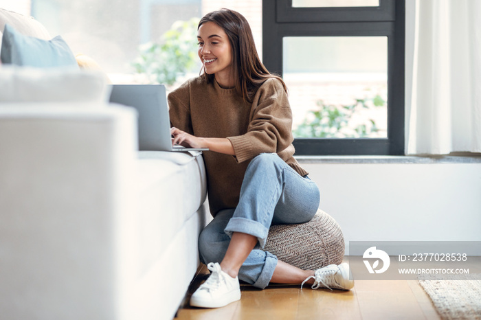 Pretty young woman working with her laptop on couch while sitting on floor in the living room at home.