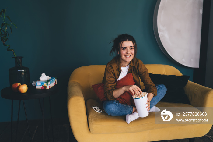 Smiling woman sitting on couch and eating popcorn while watching TV