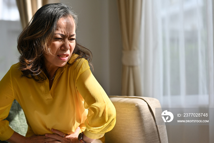 Senior woman holding belly, feeling discomfort, unhappy older female sitting on couch, suffering from stomachache, food poisoning, gastritis, abdominal pain.