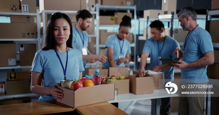 Asian beautiful young female volunteer holding box with person food supply smiling for camera. Charity help. Poor people getting aids. Multi-ethnic teamwork.