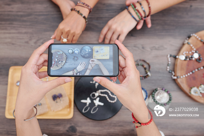 Woman photographing hands with handmade bracelets and other beautiful jewelry on wooden table