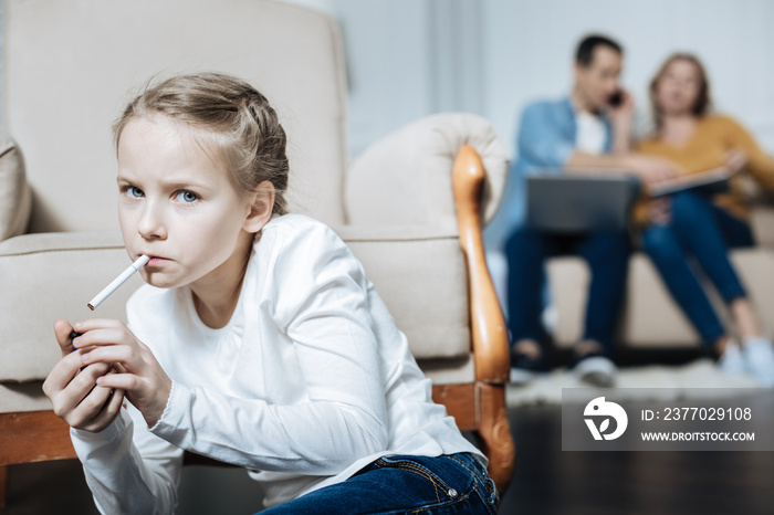 Cigarette. Poor blue-eyed little girl holding a cigarette and smoking while her parents working on their laptops