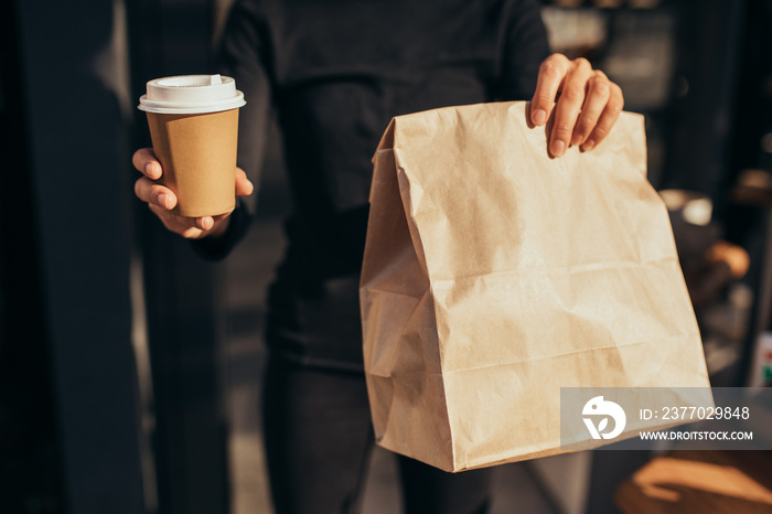 Young woman holding takeaway food and coffee to go, standing near restaurant window. Social distancing during quarantine caused pandemic.