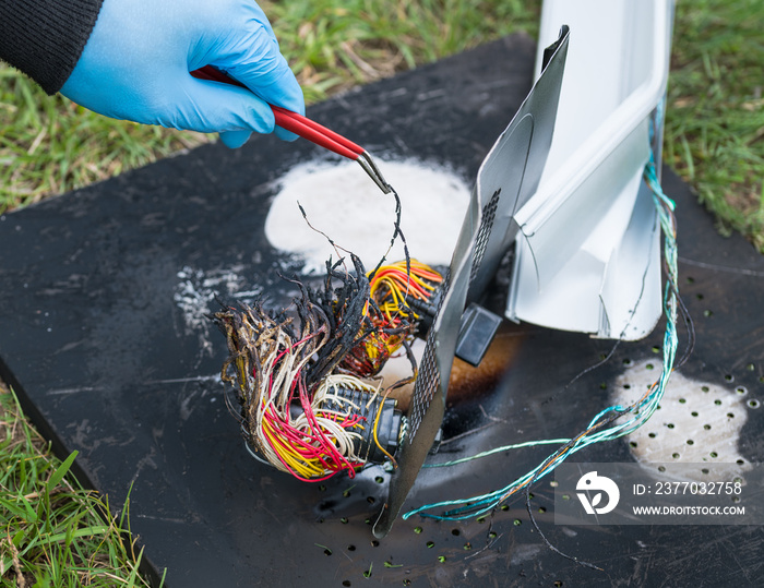 Hand in protective glove holding damaged electric wires with tweezers. Detective work on charred wreckages in grass. Investigation of air, road or railway crash. Industrial accident or terrorist act.