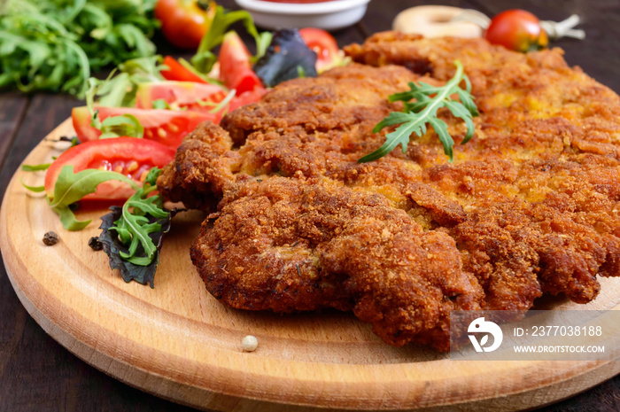 A large Viennese schnitzel  and tomato salad on a cutting board on a dark wooden background. Close up