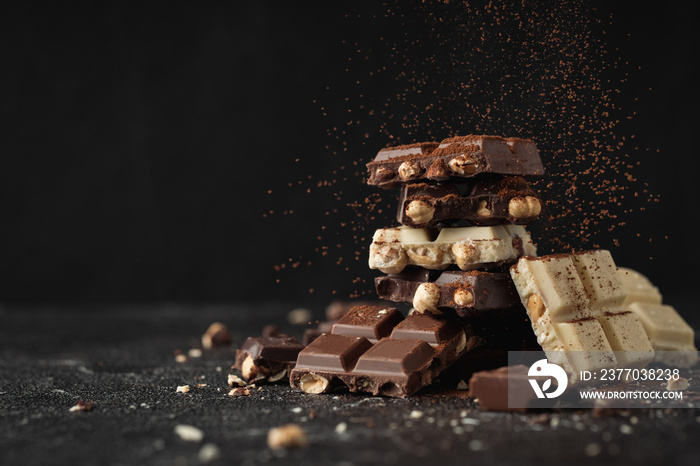 White, Milk and Dark square chocolate bars crushed with nuts on the black background and sprinkled with cocoa powder