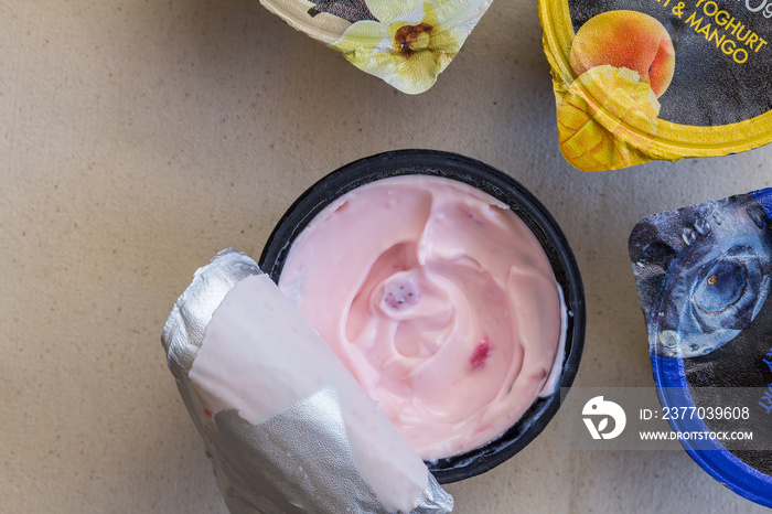 Strawberry yogurt in black plastic cup on linen background with foil lid opened and three other fruit yoghurt cups unopened