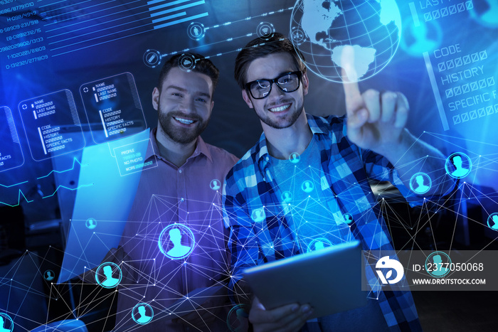 Cheerful team. Excited young clever programmers smiling while standing in front of a futuristic transparent screen with documents and gadgets in their hands