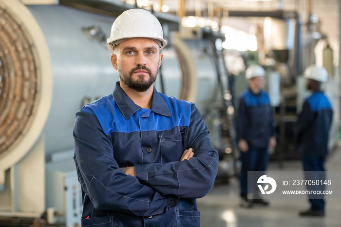 Serious young foreman in workwear and hardhat crossing his arms by chest while standing in front of camera against his two subordinates