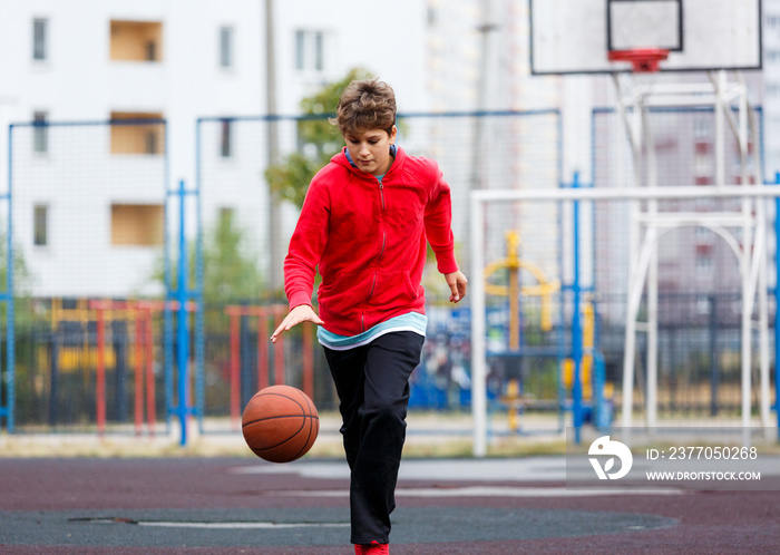 Cute boy in red t shirt plays basketball on city playground. Active teen enjoying outdoor game with orange ball. Hobby, active lifestyle, sport for kids.