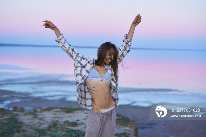 Sensual brunette perfect slender body woman posing on pink lake with naked belly at evening, raised arms. Dreaming concept, beautiful nature scenery