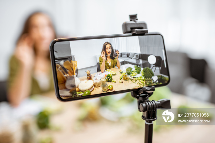 Young woman recording on a smart phone her vlog about healthy eating. Sitting at the table with lots of green vegan food ingredients at home, close-up on a phone screen