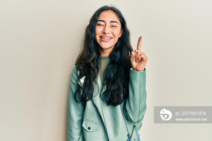 Hispanic teenager girl with dental braces wearing green leather jacket smiling with an idea or question pointing finger up with happy face, number one