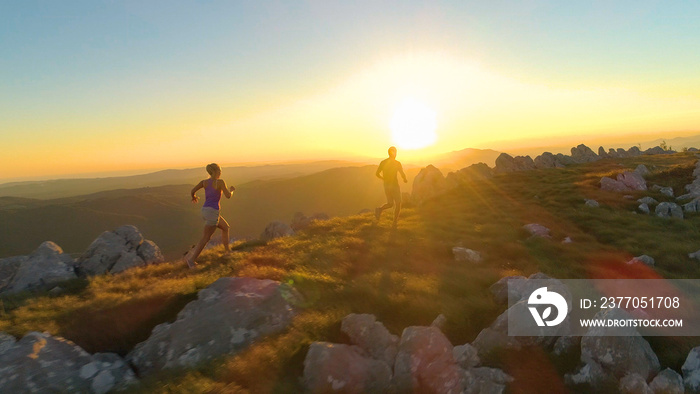 AERIAL, LENS FLARE: Athletic young couple jogging along a scenic grassy trail.