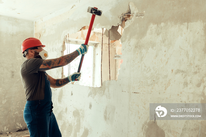 builder with a hammer in his hands breaks the cement wall The builder is dressed in a protective suit and helmet